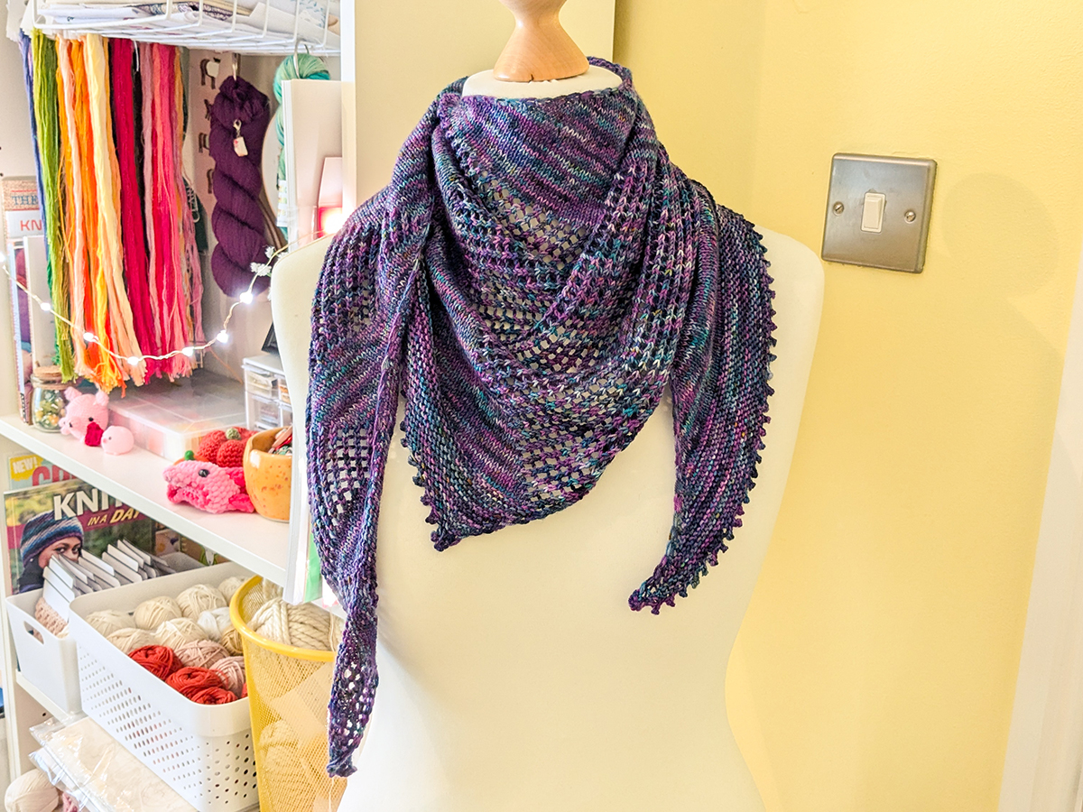 A knitted crescent shawl in variegated purple and blue yarn, worn scarf-style by a tailors dummy.