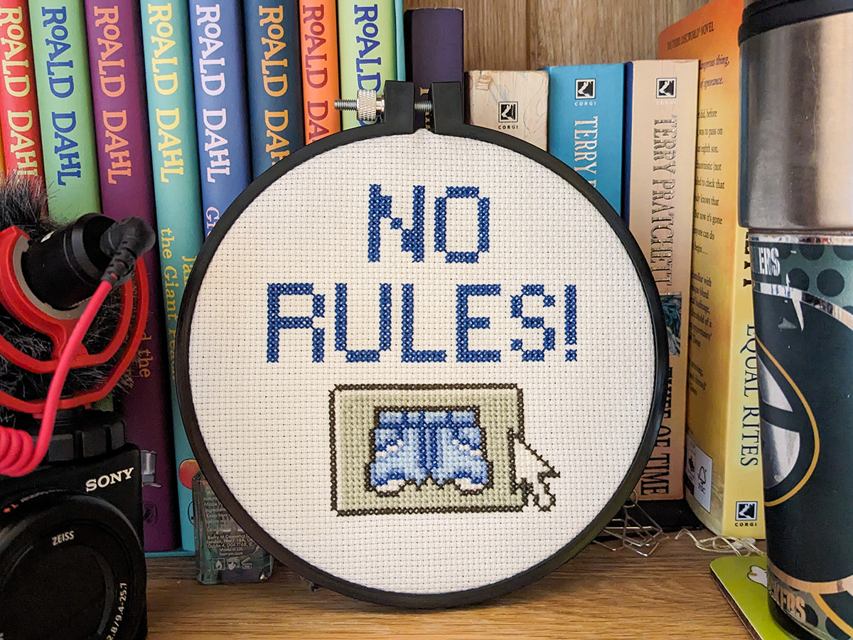 A Eurovision-themed cross stitch project in a hoop, standing on a bookshelf. It says 'NO RULES!' and features a pair of cutoff denim shorts on an old fashioned button like the ones you'd find in Windows 95.
