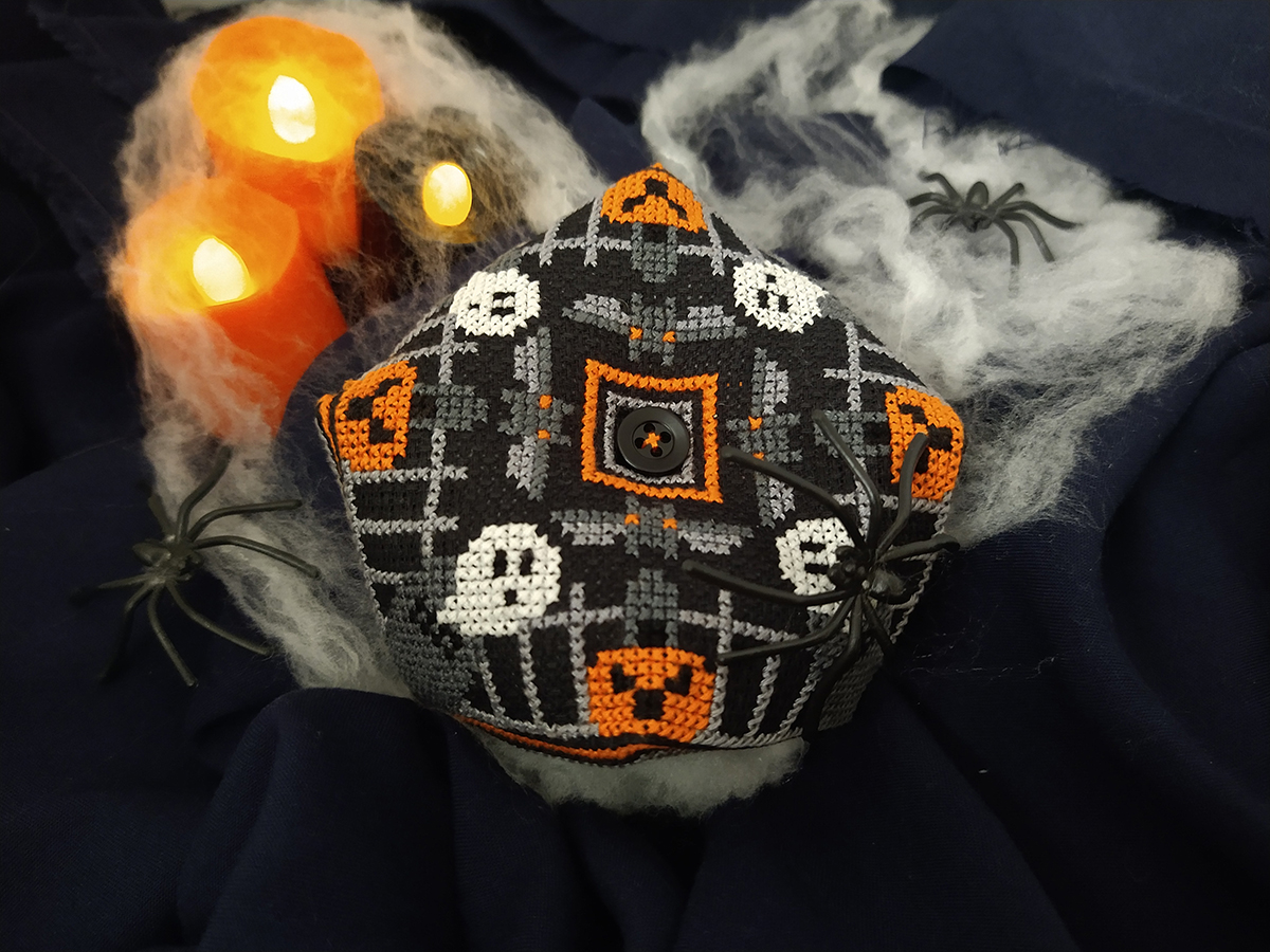 A cross-stitched Halloween biscornu in white, grey and orange on black fabric. It has a repeating pattern of pumpkins, ghosts and bats.