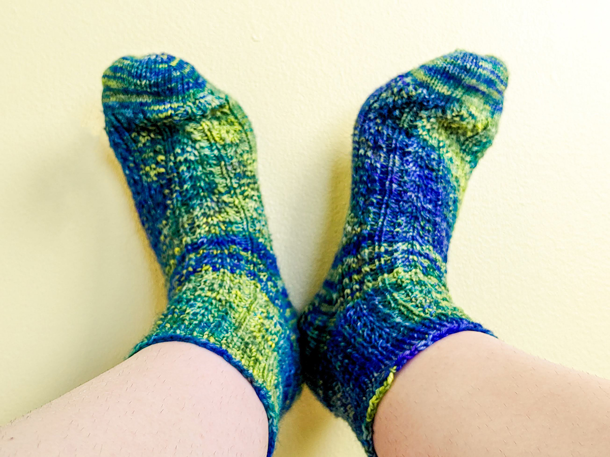 A pair of knitted socks in wildly variegated, very bright blue and green yarn that obscures the stitch pattern but it very fun.