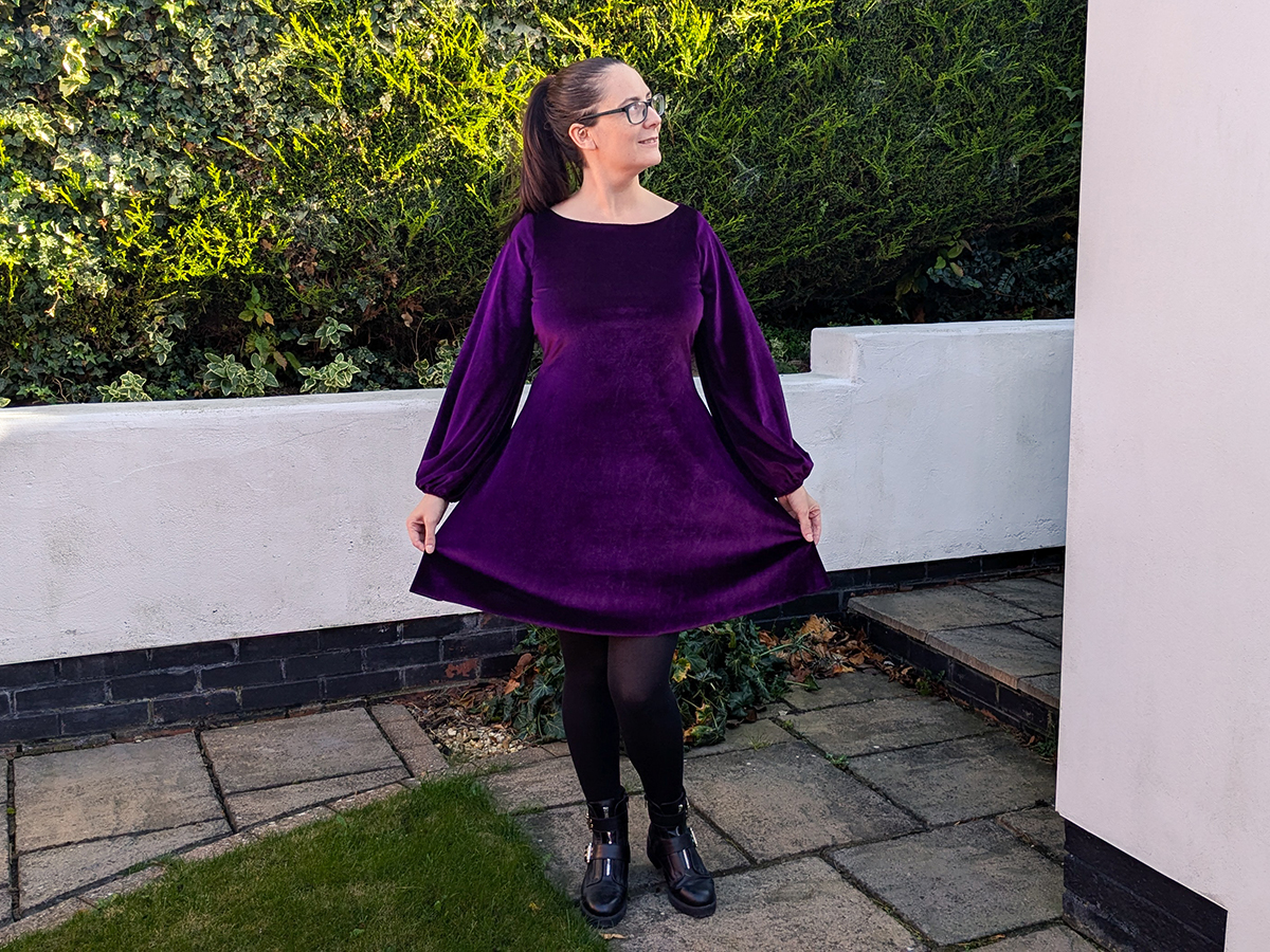 A purple velvet dress on a tailors dummy. It's knee length, with long flouncy sleeves and a boat neck.