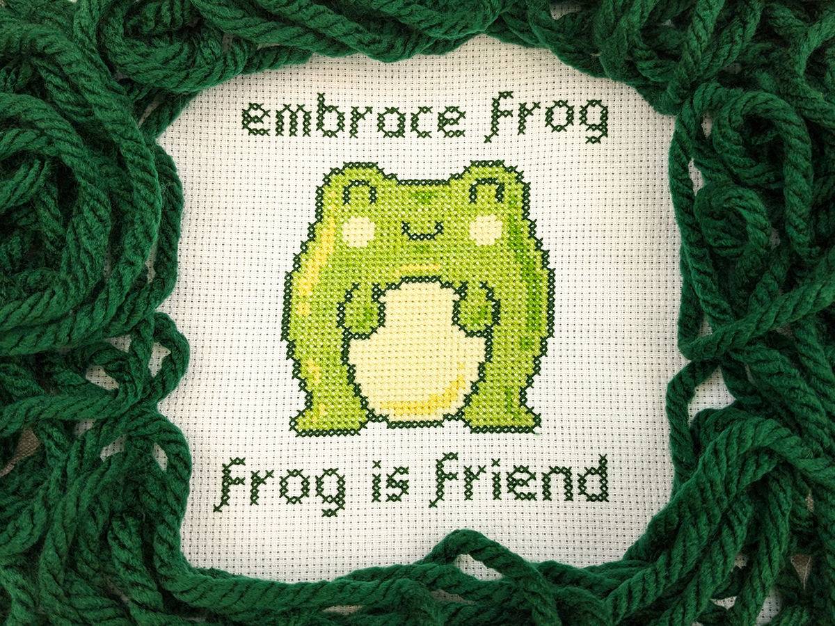 A cross stitch project with a cute cartoony frog character surrounded by the text 'embrace frog, frog is friend'.